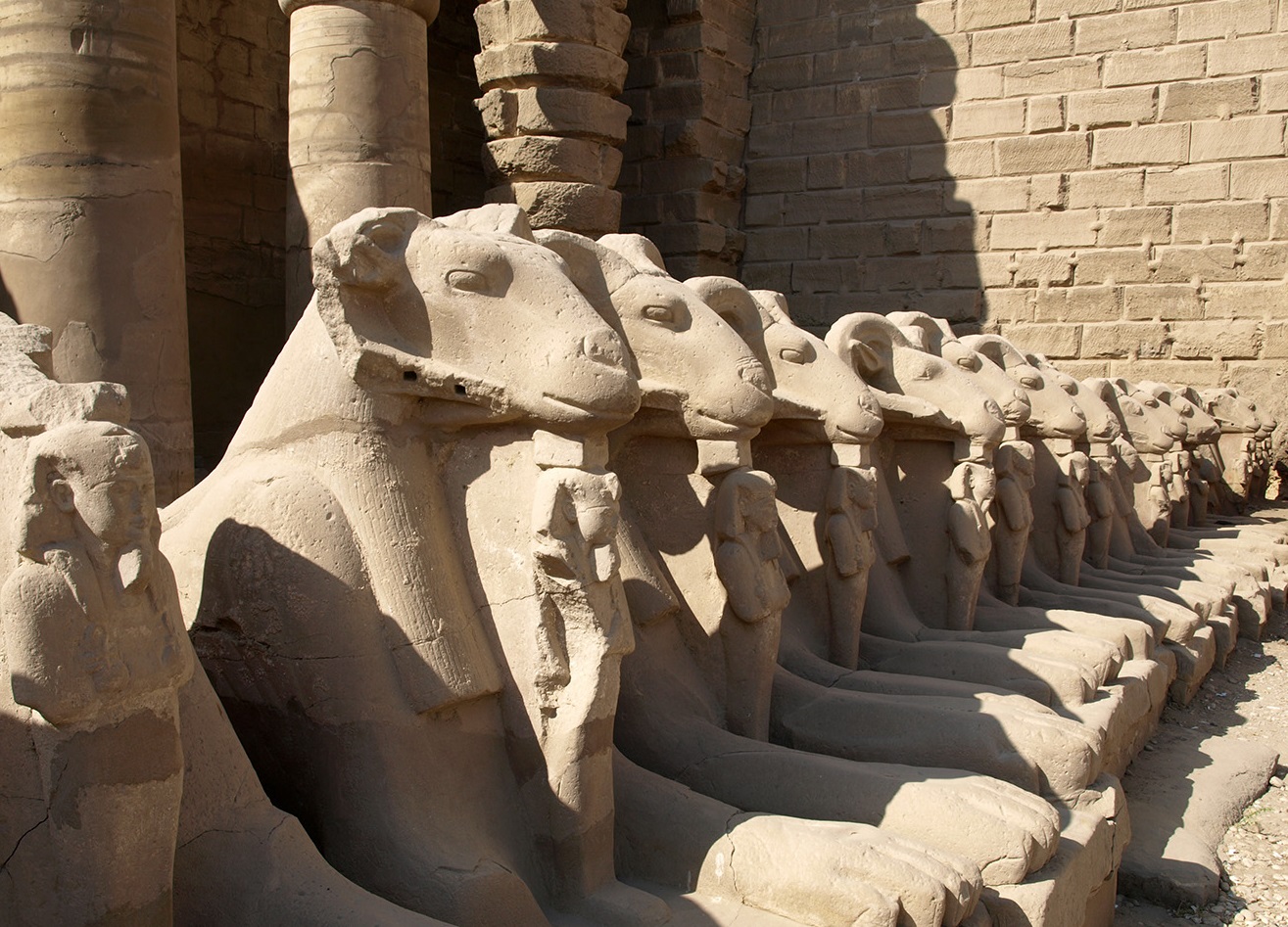 上图：建于主前2000年的古埃及卡纳克神庙（Temple of Karnak）入口的狮身公山羊像，公山羊象征古埃及最高神阿蒙-拉（Amun-Ra），站在公山羊脚前的是法老，象征最高神保护法老。多毛的公山羊是古实（Kush）的主神，埃及征服古实之后，认为古实主神就是太阳神阿蒙-拉，从此公山羊成为阿蒙-拉的化身之一。由于公山羊是多产的象征，所以公山羊形状的阿蒙神又与男性生殖神敏（Min）组合成阿蒙-敏（Amun-Min），进而演化成希腊淫乱的山羊神潘（Pan）。库努牡神（Khnum）也长着一个公羊头。